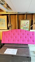Front view of pink upholstered headboard with button tufting and curved top edge, shown leaning against a wall.