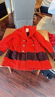 Red wool jacket with buttoned front and dark horizontal stripes, laid flat on a wooden chair.