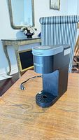 Black Keurig coffee maker machine sitting on a wooden table with chair and table in background.