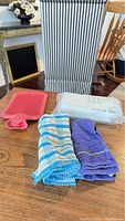View of various miscellaneous kitchen items including a striped fabric laundry hamper, red silicone hot water bottle, packaged white cleaning cloths, and two kitchen towels in blue/white and purple.