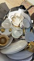Top view of a black bag containing various cups, saucers, glassware, and white serving plates showing floral and gold-trimmed designs