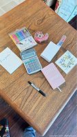 Lot of assorted office supplies arranged on a wooden table including a calculator, several notebooks, colored markers, a tape dispenser, a mini stapler, and a black pen.