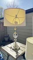 Single silver-tone lamp with clear glass sphere and white drum shade on a wooden side table outdoors