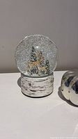 Snow globe with deer and pine trees inside, white glitter snow effect, birch-style base.