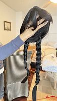 Hand holding black synthetic wig with two braided pigtails, showing smooth hair texture and style.