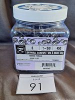 Front view of clear plastic container with white lid labeled with drywall screw details and quantity, placed behind lot number card 91.