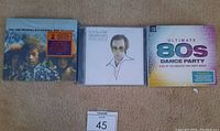 Photo of three unopened music CDs laid out on carpet showing The Jimi Hendrix Experience box set, Elton John's Greatest Hits, and Ultimate 80's Dance Party compilation.