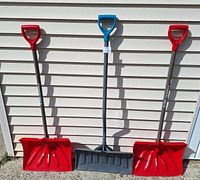Photo showing three snow removal tools: two red snow shovels on either side and one black snow pusher with a blue handle in the middle, all positioned against a light-colored vinyl siding wall.