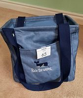 Blue-grey tote bag with navy straps and front pocket showing whale embroidery and 'Ride the waves' text on carpeted floor against green wall.