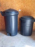 View of two black plastic trash cans against plywood wall, showing overall condition and size difference
