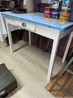 Full view of antique kitchen table showing porcelain top with blue painted design, central drawer with metal handle, and white wooden legs and frame.