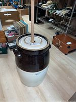 Full view of large pottery butter churn with wooden plunger inserted through the lid, visible kitchen background.