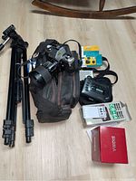 Full view of lot showing Minolta X-370 camera on a camera carry case, adjustable tripod, boxed Blacks CB 64 flash, boxed Sigma zoom lens, and Sony Walkman cassette player along with small transistor radio and single use camera in packaging.