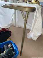 Side view of folding card table showing metal leg and frame with brown vinyl top, some items around the base not included in lot.