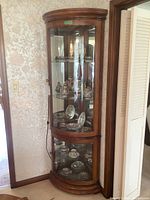 Full view of the tall curio cabinet placed in a corner, showing its curved front glass panel, wood finish, and mirrored sides with glass shelves inside containing decorative items (not included).