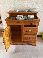 Front view of knotty pine wooden dry sink with open cabinet door and drawers, dishes on upper shelf