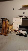 Photo displaying wooden easels next to canvas boards, briefcase, and various art and upholstery supplies on the basement floor.