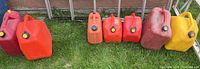 Seven plastic gas cans lined up on grass near a shed, showing different colors and sizes.