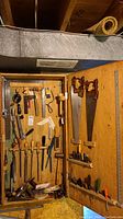 Wide view showing the wooden cabinet with multiple tools mounted inside, including saws, hammers, measuring rulers, pliers, files, chisels, and screwdrivers.