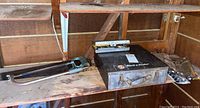 Photo of metal punch tool, Black & Decker impact wrench metal case, and hand saws on workbench in shed.