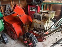 Side angled view of the orange Ariens Sno-Thro snowblower showing engine, tires with chains, chute, and rust.