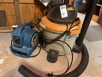 Photo shows 9 gallon shop vacuum with grey and orange body, black hose and attachments, next to blue XPower air mover fan with power cords visible.