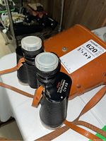 Front angle of black ARL Wetzlar binoculars with white lens caps and brown leather case behind