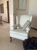 Front view of the cream-colored wing chair with the decorative pillow and arm covers visible.