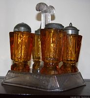 Five-piece amber mitred diamond pattern cruet set on clear pressed glass holder, including four shakers/cruets with metal lids and one clear glass holder with central handle