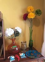 Overall lot of art glass vases, plaster fish, glass chili pepper, and decorative plate on a wooden surface and against a yellow wall.