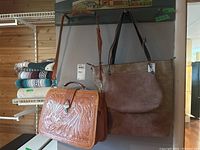 Three bags hanging on a wall hook, including a tooled leather satchel and two brown faux suede bags, one crossbody and one tote style