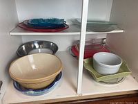 Wide shot of cabinet shelving showing multiple bakeware and serving dishes, large beige mixing bowl, glass dishes in various colors and shapes