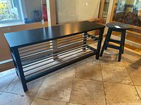 Photo showing the black laminated bench with a table top and metal bars for lower shelving and a round wooden stool adjacent to it on tiled floor.