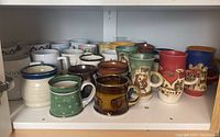Wide view of shelf containing various ceramic mugs in different colors, designs, and styles including Godiva mugs and artist signed pieces.