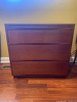 Front view of the brown wooden chest of drawers showing three smooth drawer fronts and simple trim detail above the top drawer.