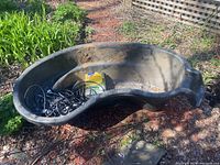 Top view of black plastic garden pond shaped irregular kidney with electric pump cables and box inside.