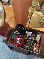 Photo showing box containing vintage wooden golf club, golf balls in various conditions including some in boxes, and golf accessories including a headcover and a green brush.