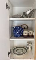 Kitchen cupboard interior with blue and white teapot, steel gravy boat, and ironstone plates and mugs on shelves
