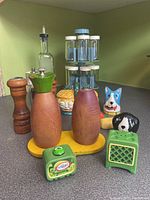 Photo showing wooden salt and pepper shakers, dog-themed ceramic shakers, green ceramic retro TV shakers, a glass oil bottle, and spice rack with jars in background on kitchen counter.