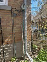 Photo showing two shepherd's hooks side by side against a brick wall, with a birdhouse mounted on a metal pole next to them in an outdoor garden area.