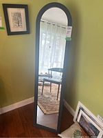 Front view of the full-length standing mirror showing its arched top and black composite frame against a green wall.