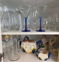 Assorted clear glasses including martini glasses with blue stems, tall glasses, shot glasses, and small tumblers on upper shelves with ceramic salt and pepper shakers and teapot on bottom shelf.