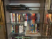 Photo of a black JVC HR-A59OU VHS player on a shelf above several VHS tapes and DVDs.