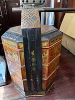 Front view of one lacquered octagonal banquet basket showing gilt scenes and wicker handle clasp and pole end