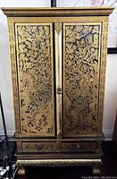 Front view of cabinet showing two doors with gold leaf foliage and bird motifs, two drawers, and raised platform base.