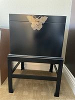 First deed box on stand showing overall black finish and brass lock, standing on carpeted floor