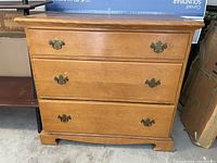 Front view of vintage midcentury 3-drawer wooden dresser showing natural wood finish and brass-tone drawer pull handles. Scratches and marks visible on surface.