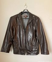 Front view of dark brown leather jacket hanging on a black plastic hanger against beige wall