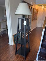 Full view of the modern black wooden floor lamp with three square shelves and a white fabric shade.