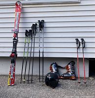 Full set of ski equipment displayed outdoors against siding. Includes red and white Atomic skis, multiple pairs of ski poles, two pairs of ski boots, and a black helmet.
